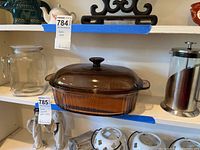 Photo showing Vision amber glass casserole dish with lid, glass pitcher, and French press coffee maker on white shelves.