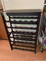 Front view of a wooden wine rack with six shelves for wine bottles on a wooden floor near a door.