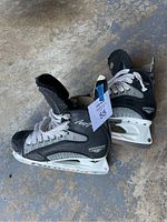 Oblique view of two black and gray Mission Amp 7 hockey skates with white laces and blade holders on concrete floor.