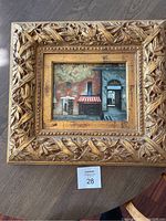 Framed oil painting of city street scene with ornate gold frame on wood floor.