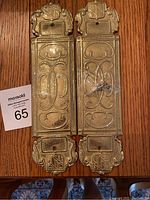 Front view of pair of brass door plates on wooden surface, showing decorative embossed details and mounting holes.