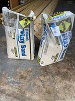 Two unopened 50 lb bags of Natural Play Sand placed on a concrete floor, with some wooden planks in the background.