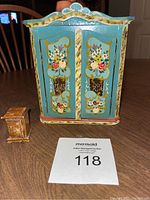 Front view of vintage painted wood doll wardrobe with floral folk art designs and small wooden accessory piece on wooden table.