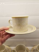 Close-up of one cream textured Lambethware cup with embossed floral pattern, shown on matching saucer and held by hand for scale.