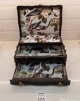 Open black wooden jewelry box showing interior bird-pattern velvet fabric lining with pin clasps for hanging jewelry, two drawers open