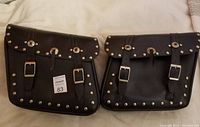 Pair of black leather motorcycle saddle bags with silver rivet accents and buckle closures laid flat on a white surface.