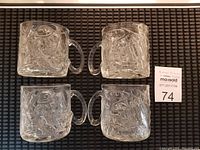 Four clear glass mugs with embossed Riddler character from Batman Forever, handles visible, laid flat on dark surface.