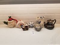 Seven miniature teapots of various designs and sizes on counter with white subway tile background, showing the variety in styles and conditions.