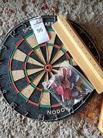 Photo of the dartboard with wood stand and a plastic bag containing darts with various flights laid on top.