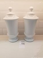 Pair of tall white ceramic apothecary canisters positioned side by side on a white counter with tiled backsplash.