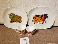 Two rectangular vintage English ironstone plates with colorful stylized beef cow illustrations and two vintage floral design ceramic mugs shown on a soft-texture background