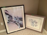 Two framed prints leaning against a wall: left is a large abstract print with black frame, right is a smaller floral print with lighter frame.