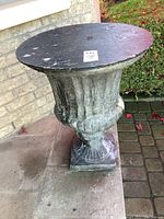 Full view of the cement planter showing urn shape and the removable black wooden top on a stone floor outdoor setting.