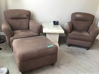 Pair of brown fabric finish chairs with visible wear, including an ottoman and a white side table with a phone on it in a living room setting.