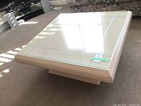 Square wood coffee table with clear glass top, beige tone, pedestal style base on carpeted floor.