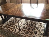 Top view of the rectangular dining table with a polished inlaid wood grain surface and a visible seam where the table halves meet. Part of a floral-pattern rug underneath the table is visible.
