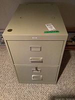 Full front view of beige metal two drawer filing cabinet, top drawer unlocked and bottom drawer featuring a key lock with key inserted.
