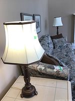 Table lamp with dark metal base and cream fabric shade, positioned on a side table next to a floral couch and wall art.