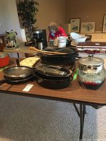 Kitchen cookware and utensils including frying pans, muffin tins, wooden spoons, glass container, and baking racks arranged on table.