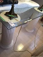 Glass top console table with beveled edges supported by two white fluted column-style metal stands, one view showing side and front angle with lamps on top (lamps not included).