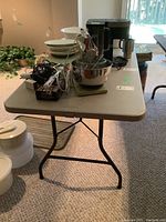 Rectangular beige folding table with black metal folding legs. The table is shown with various kitchen items on top but contents are not included.