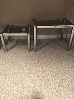 Two mid-century modern style side tables with tubular chrome legs and smoked glass tops shown from front, on carpet in indoor setting.