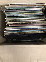 Top down photo of approximately 60 vinyl records in a cardboard box, showing varied album cover edges.