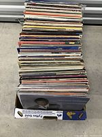 Stack of over 50 vinyl records in a box, showing the side edges of record covers.