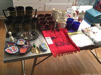 Wide shot showing entire lot laid out on table with mugs, cups, patches, pins, plates, towel, and trophy
