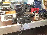 Photo showing Pentax PC35AF-M camera with manual, Polaroid One Step camera, Minolta XL401 movie camera, camera cases, miniature transistor radio, and other radios and headphones.