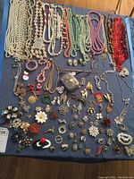 Overview of assorted costume jewelry pieces laid out on a blue cloth, includes necklaces, earrings, pins, and a metal trinket box