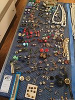 Wide view of many pairs of earrings in varied colors and styles, several necklaces, and watch visible on a blue perforated surface.