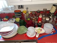 Overview of various vintage kitchenware items including stacked colorful bowls, white dishes, glass bottles, and kitchen utensils on a kitchen counter.