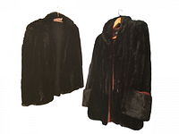 Photo showing two black sheared mink fur coats hanging on hangers, one with wide cuffs and collar, the other simpler.