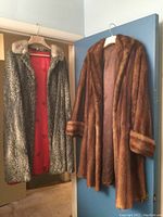 Two fur coats hanging, lamb mouton cape on left with grey textured fur and red lining, mink coat on right with brown fur and cuffs