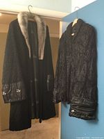 Photo showing two black lamb mouton jackets hanging side by side. The jacket on the left has a light fur collar and leather cuffs with buttons, the other jacket has a swirled texture.