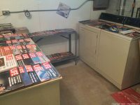 Photo showing several stacks of Life magazines with colorful covers and visible text, arranged on tables and shelves in a basement room with washer/dryer.