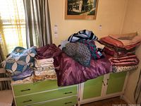 Photo of mixed stacked linens including sheets, blankets, knitted sweaters, and a purple comforter with matching pillowcases on green furniture.