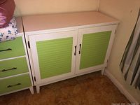 Front view of the Mid Century Modern wood cabinet showing white frame and green shutter-style doors with black handles, placed next to a matching green drawer unit.