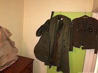Green military trench coat, Army ROTC jacket with patches, and military bags hanging on a wall