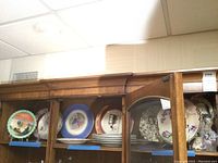 Photo showing multiple decorative plates on wooden shelves including a mix of floral, blue and gold accented plates and some stacked plates.