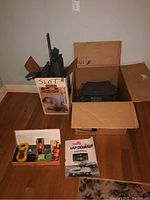 Two boxes containing black plastic slot car track pieces, one cardboard box showing Repco branding, on wood floor near wall corner.