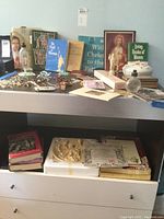 Overview photo of various religious books, rosaries laid on the table, holy water bottle, framed images, bowl, crucifixes, and pins.