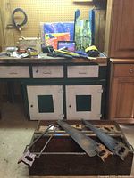 Hand saws leaning against a wooden crate with tools on a green and white workbench. Various tool boxes, packaging, and blueprints hanging on wall pegboard.