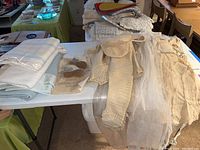 Vintage clothing and textiles laid out on a table including folded blankets, knitted baby outfit, baby shoes, and gowns.