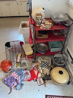 Photo showing assortment of vintage kitchenware on and around red utility cart including pots, pans, aprons, mitts, recipe booklets, baby bottles.