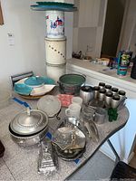 Photo of vintage kitchenware items including aluminum serving bowls, trays, spice rack with canisters, Fire King cups, glass dishes, and ceramic plates on kitchen counters