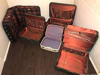 Five vintage suitcases arranged on the floor showing interiors and exterior patterns; three green suitcases, one plaid garment bag, one white suitcase.