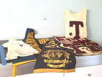 Overview of vintage school memorabilia items laid out on a metal surface, including Towson High School pennants in maroon and cream, a navy and yellow Johns Hopkins pennant, and a cream knit sweater with a large maroon letter 'T' and star emblem.
