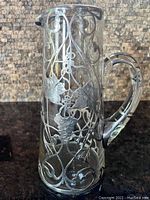 Front view showing tall cylindrical glass body with silver overlay vine and grape decoration